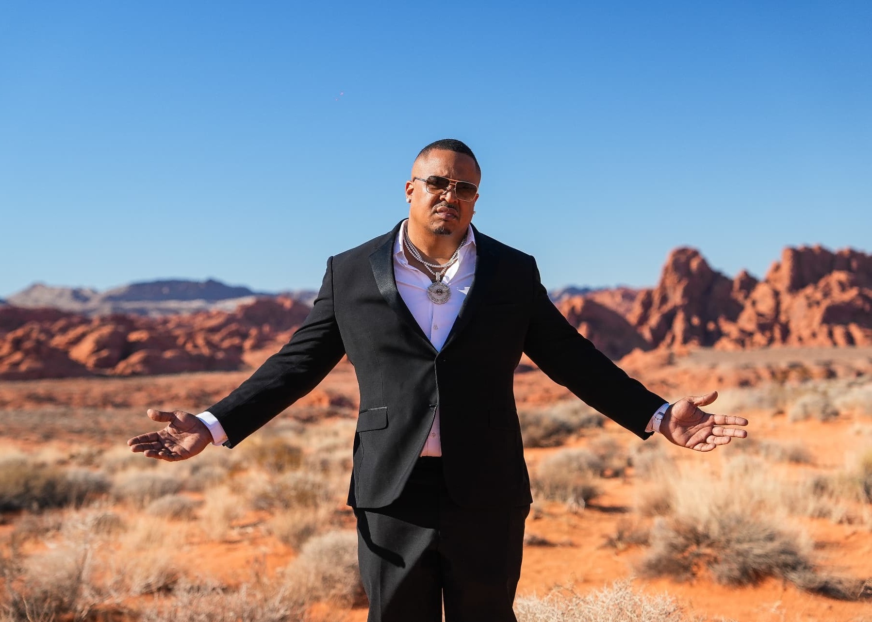 Chef Geoff Cole standing in a red-sand desert with sandstone ridges behind him, wearing a black suit jacket, white open-collar shirt, diamond chains and gold-tinted aviators, arms open at his sides