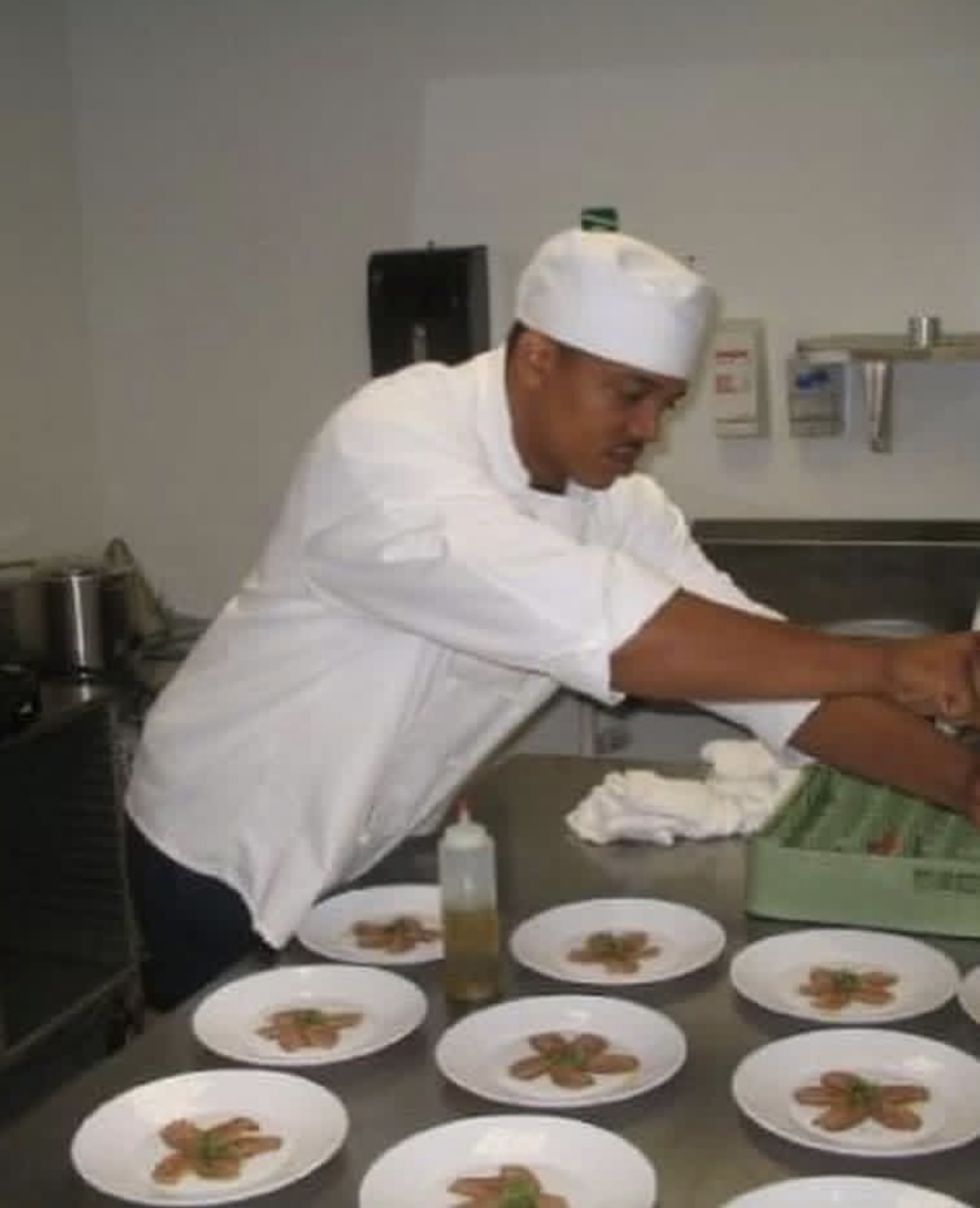 Geoff Cole in a white chef coat and hat, plating dishes at a stainless steel prep table