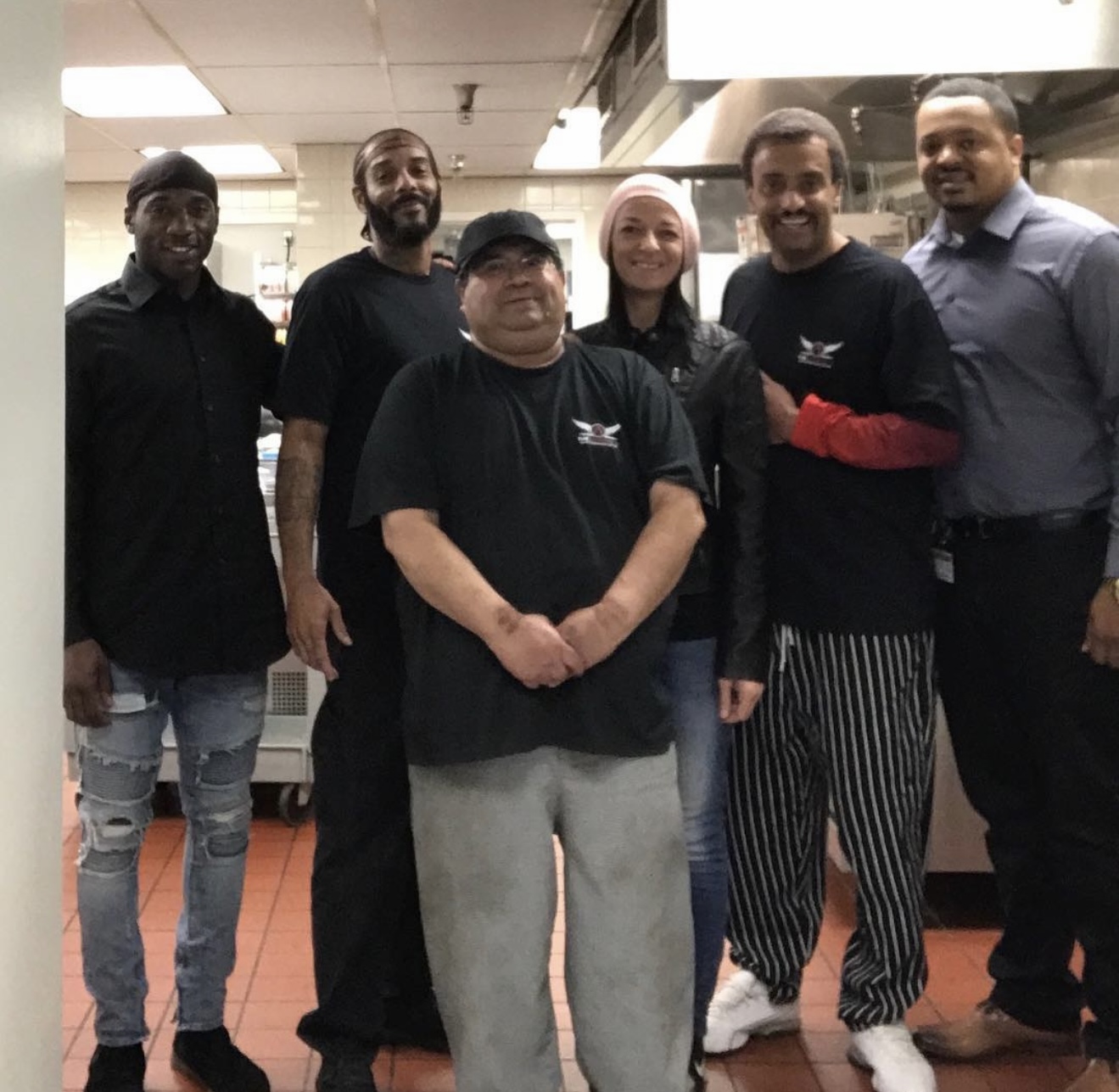 A team of six in black The Admiral's Experience shirts standing together in a commercial kitchen