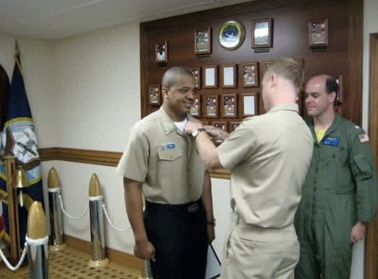 An officer in a tan uniform pinning an insignia onto a collar in front of a wooden plaque board