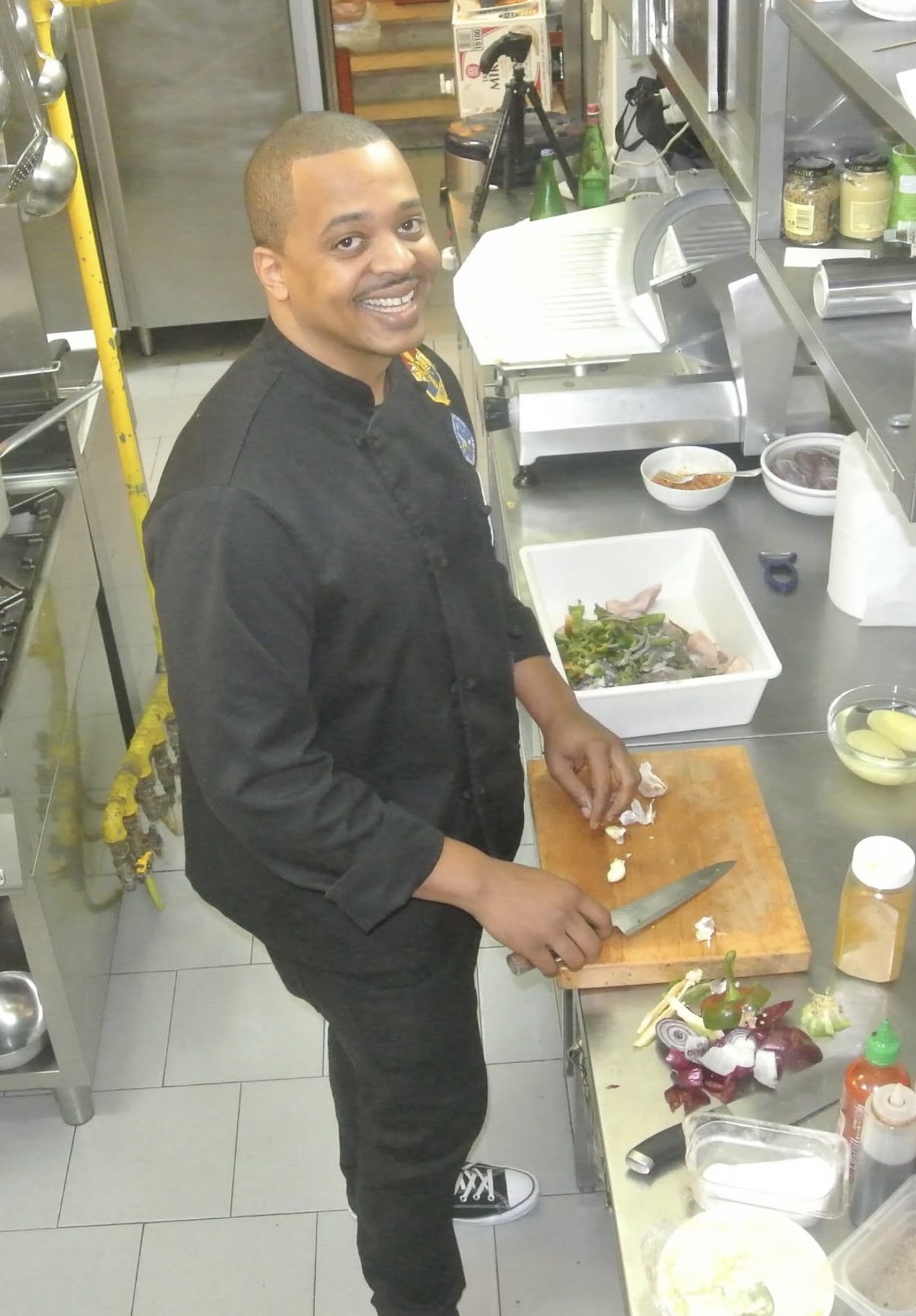 Geoff Cole in a black chef coat, smiling as he cuts vegetables on a wooden board in a commercial kitchen