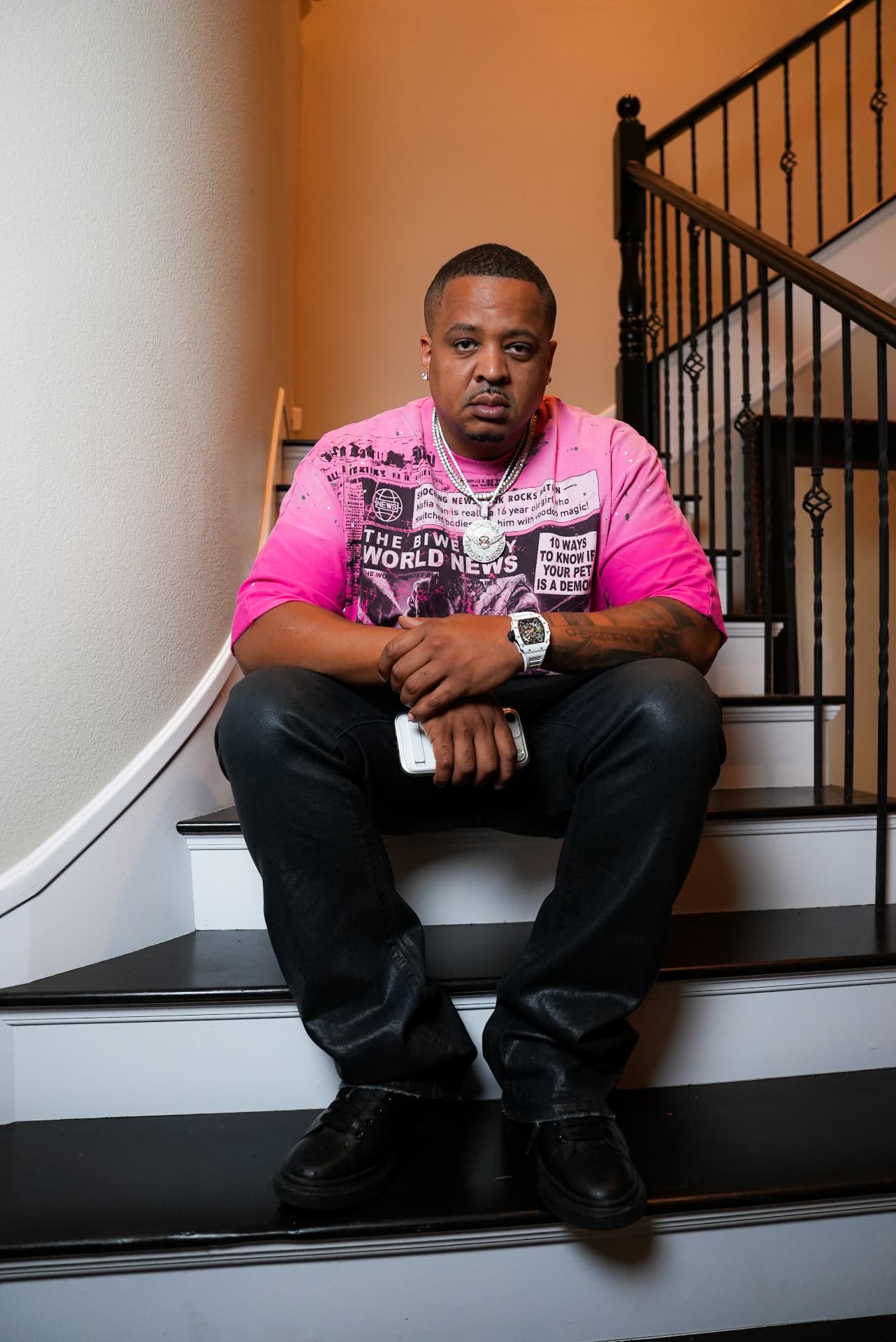 Geoff Cole in a pink shirt and chains seated on a staircase, arms resting on his knees