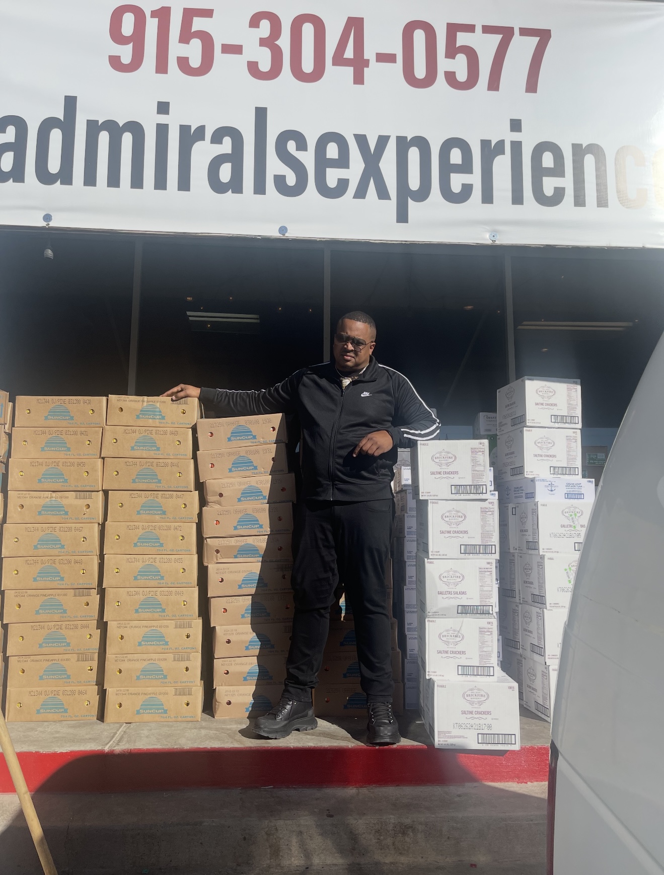 Chef Geoff Cole standing beside tall stacks of supply boxes at a loading dock in a black track jacket, sunglasses and gold chain