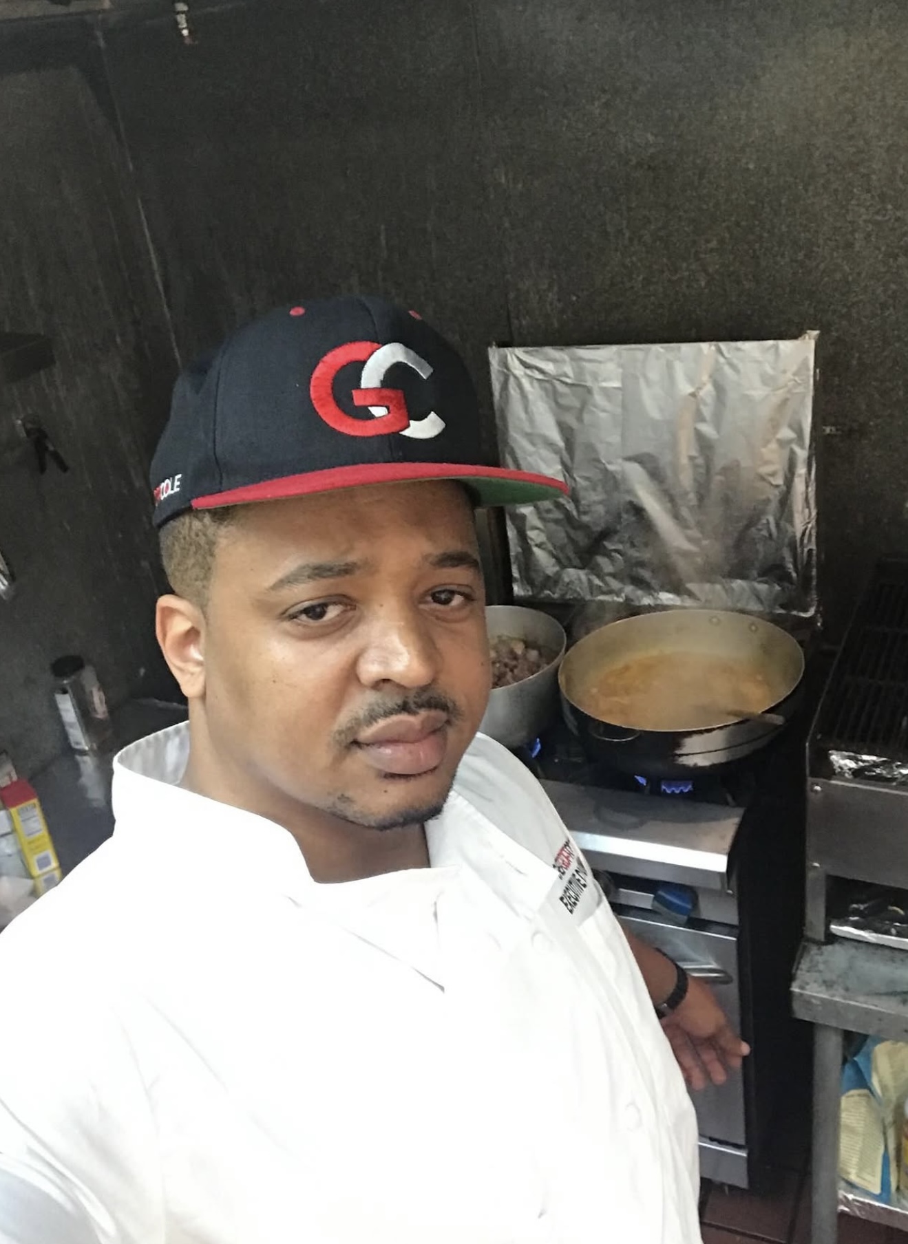 Geoff Cole in a chef coat and a GC cap, photographing himself in front of a simmering stove line
