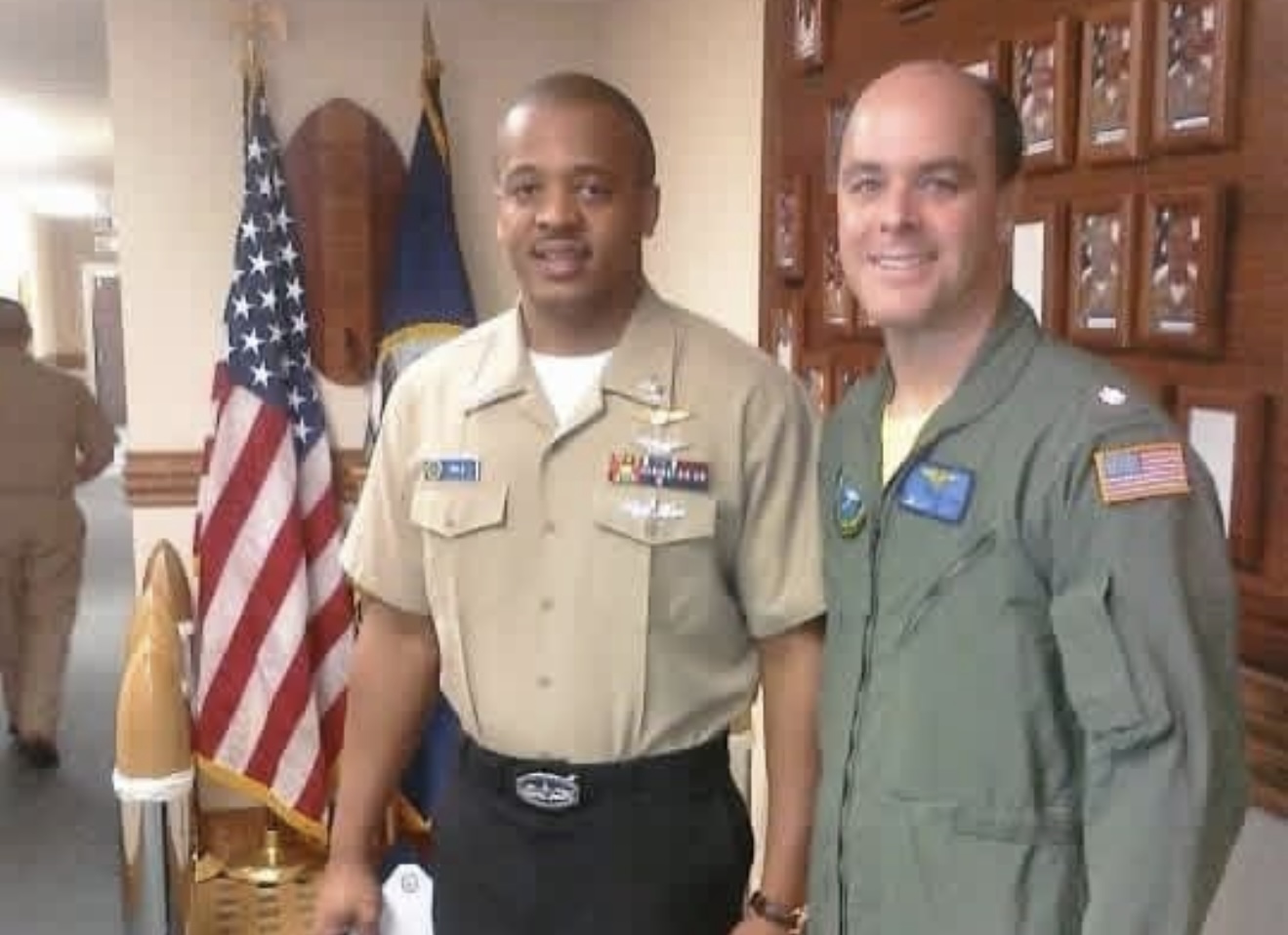 Two men smiling beside an American flag — one in a tan uniform shirt with ribbons, the other in an olive flight suit