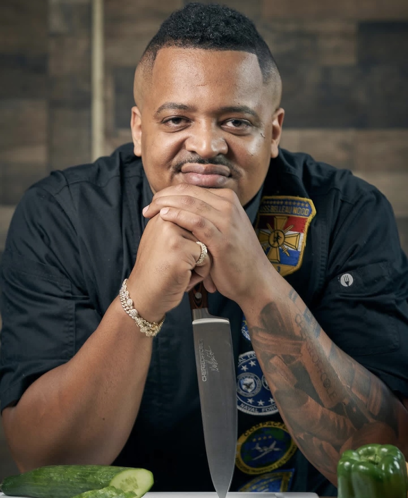 Geoff Cole in a black chef coat covered with unit patches including USS Belleau Wood, chin resting on his clasped hands behind an upright chef's knife engraved ChefGeoffCole, sliced vegetables in front