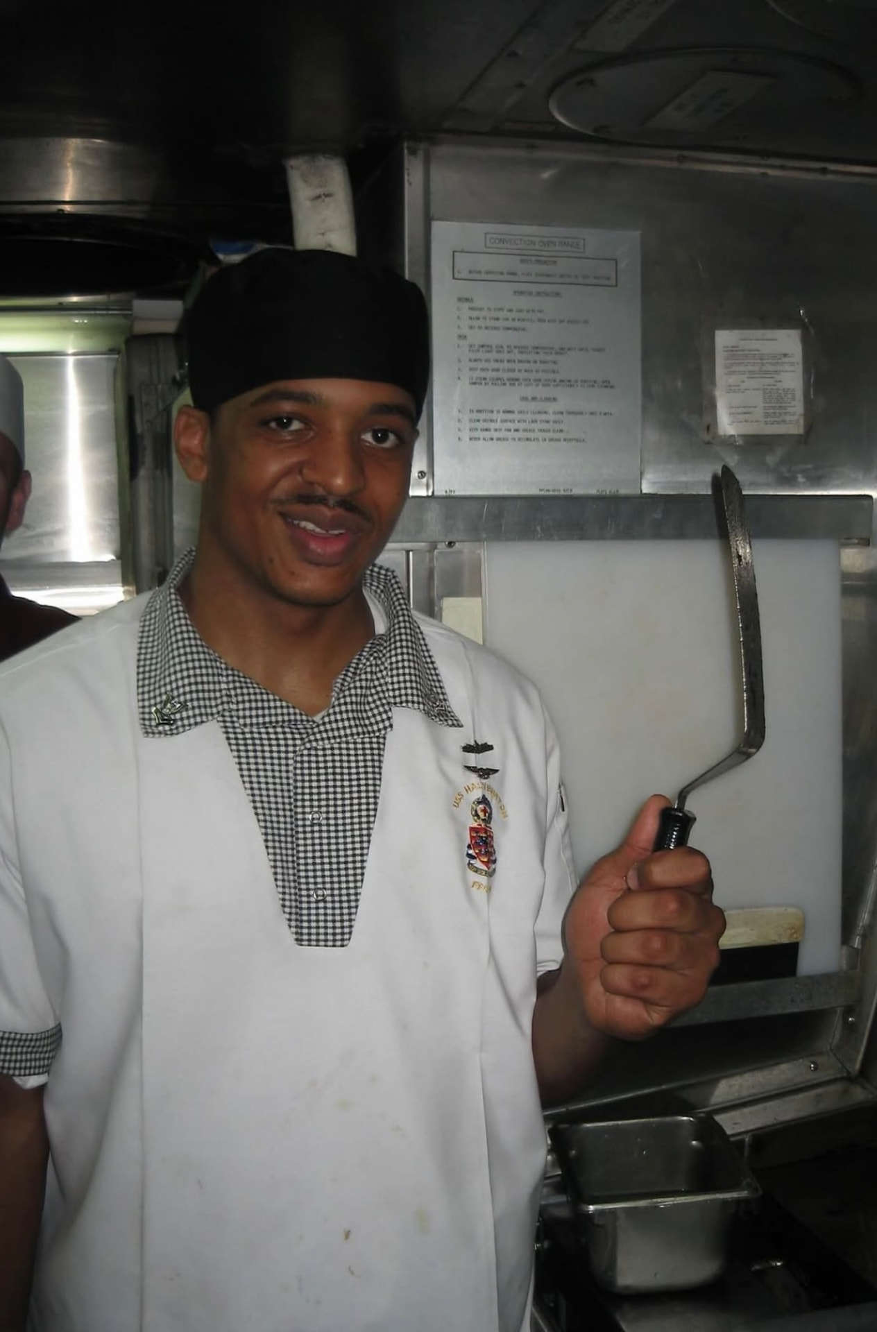 Geoff Cole in a white Navy chef coat with a USS Halyburton crest, holding a spatula in a ship's galley