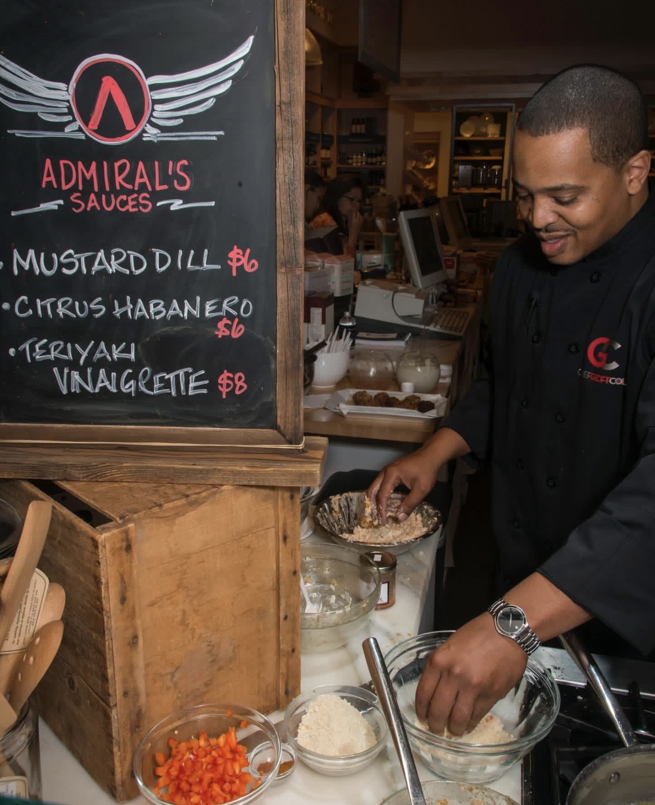 Geoff Cole in a black chef coat mixing ingredients beside an Admiral's Sauces chalkboard menu at a retail counter