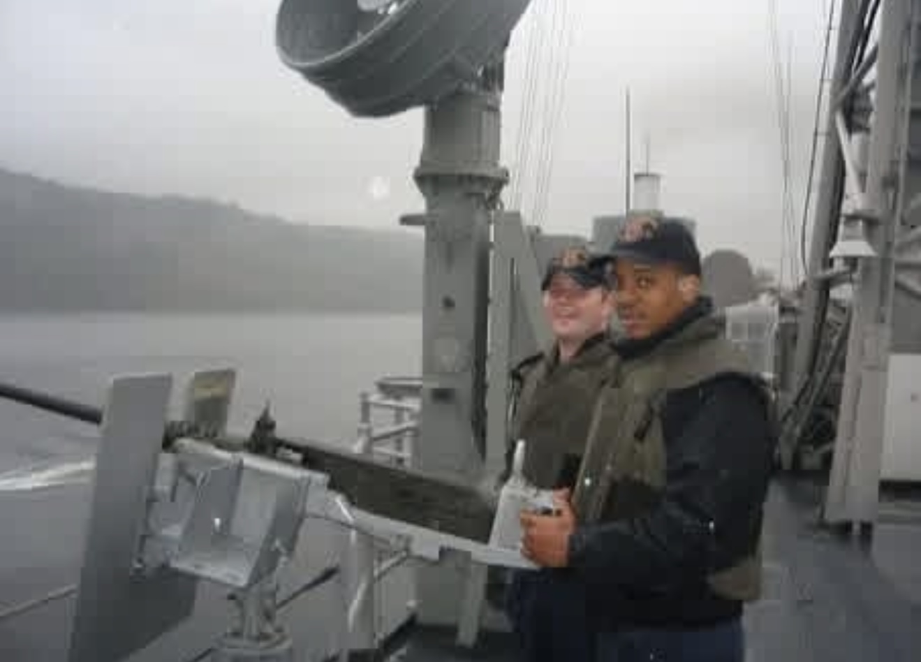 Two men in tactical vests and ball caps standing beside a mounted deck gun aboard a naval vessel