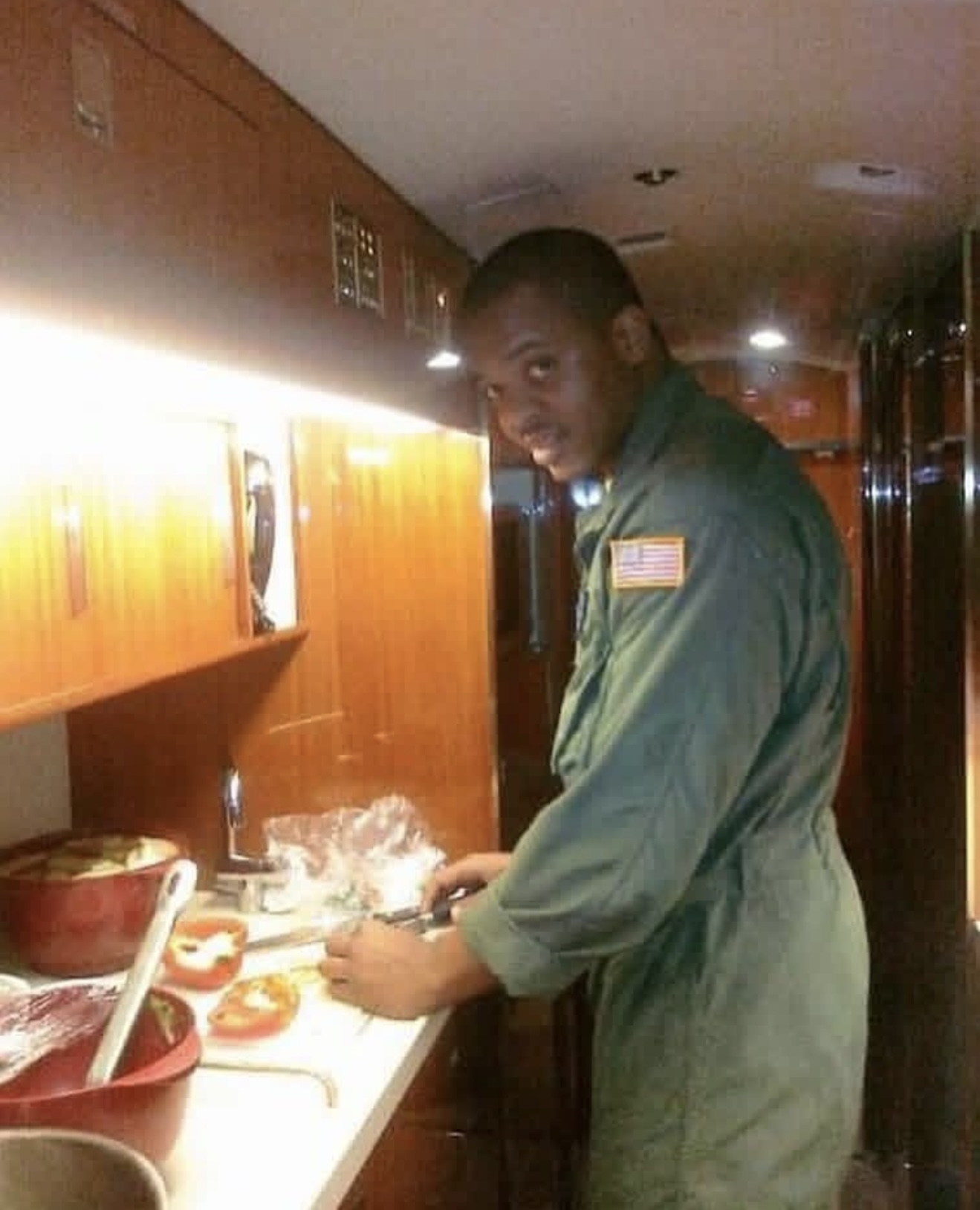 Geoff Cole in an olive flight suit with a U.S. flag patch, slicing vegetables in a wood-paneled galley