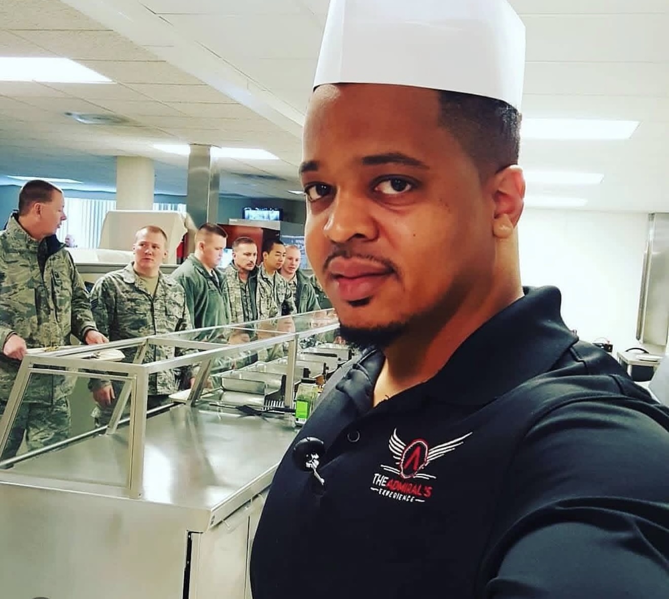 Geoff Cole in a white chef hat and a black The Admiral's Experience polo, taking a selfie at a serving line with service members queuing behind