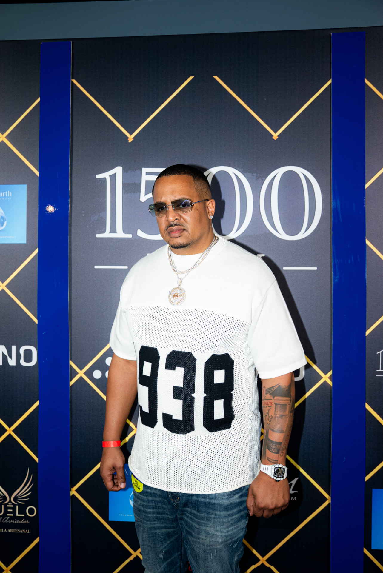 Chef Geoff Cole at a red-carpet event in a white number 938 mesh shirt, sunglasses and diamond chain, in front of a gold-and-black step-and-repeat backdrop