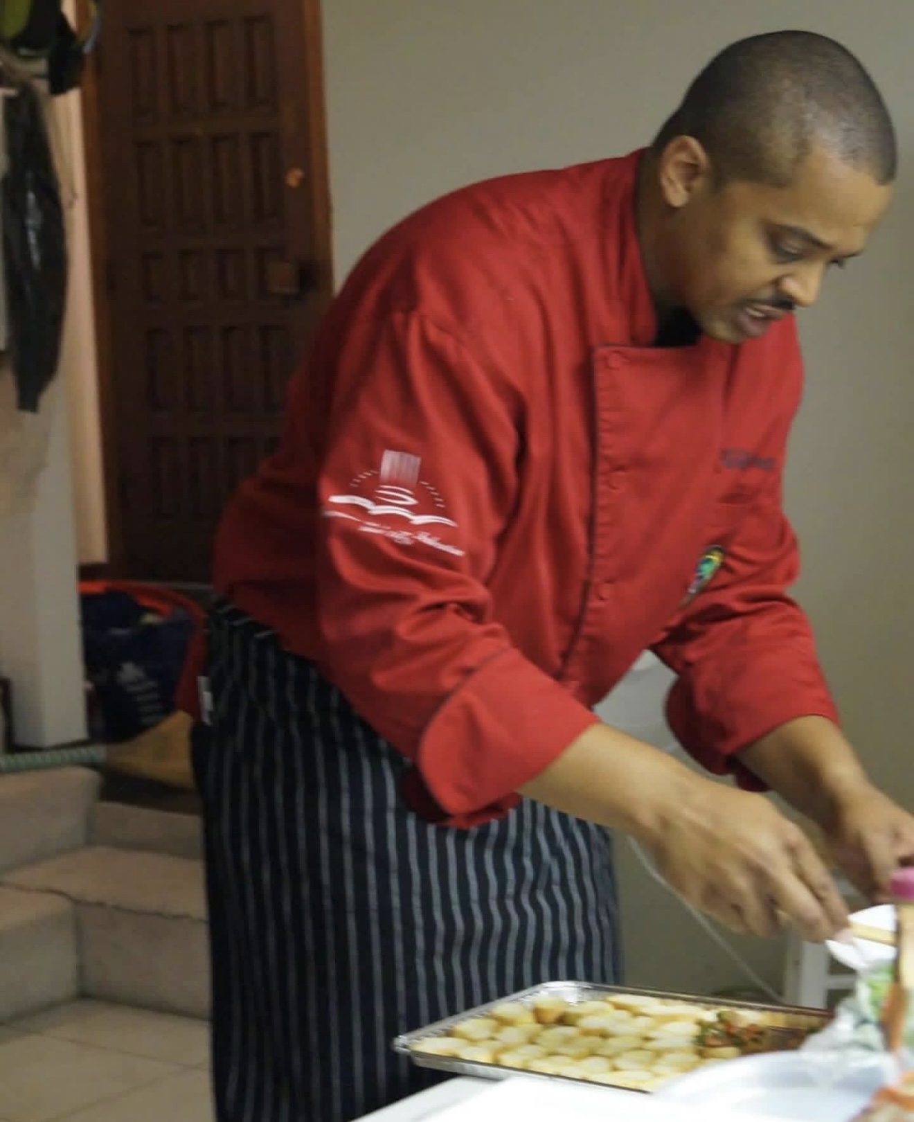 A man in a red chef coat and black-and-white striped apron, leaning over a tray of sliced bread appetizers