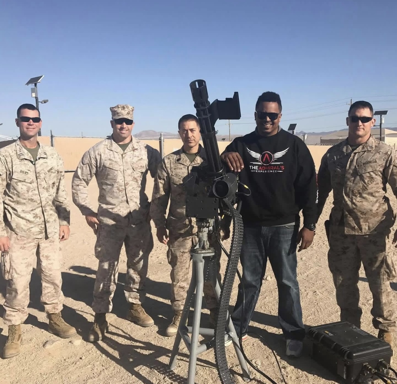 Geoff Cole in a black The Admiral's Experience sweatshirt standing with U.S. Marines beside a mounted weapon on a desert range