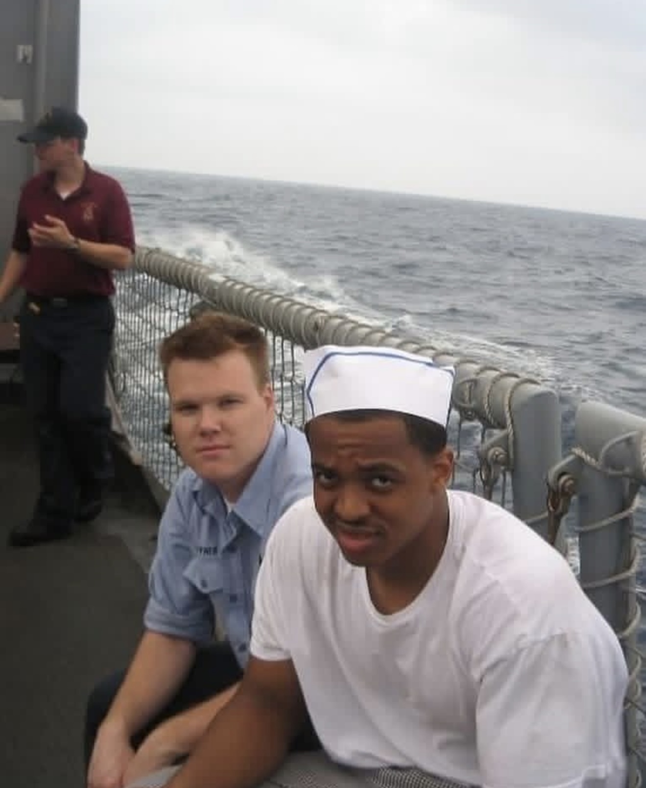 A man in a white t-shirt and white garrison cap seated on a ship's deck, open ocean behind him